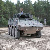 Der Radschützenpanzers Schakal steht auf einem schlammigen Platz vor einem Nadelwald.