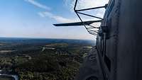 Aus dem Seitenfenster eines Hubschraubers sieht man eine hügelige bewaldete Landschaft.
