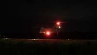 Ein Hubschrauber mit roten Positionslichtern hebt im Dunkeln von einem Flugfeld ab.