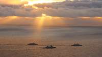 Drei Kriegsschiffe fahren auf dem Meer bei tiefstehender Sonne vor bewölktem Himmel.