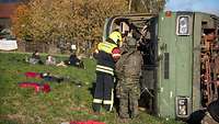 Ein umgekippter Bus auf einer Wiese, davor ein Feuerwehrmann und ein Soldat, daneben Verletzte.
