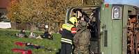 Ein umgekippter Bus auf einer Wiese, davor ein Feuerwehrmann und ein Soldat, daneben Verletzte.