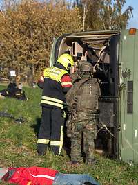 Ein umgekippter Bus auf einer Wiese, davor ein Feuerwehrmann und ein Soldat, daneben Verletzte.