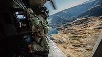 Ein Soldat kniet an der offenen Tür eines Hubschraubers, der über eine Gebirgslandschaft fliegt.