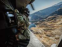 Ein Soldat kniet an der offenen Tür eines Hubschraubers, der über eine Gebirgslandschaft fliegt.