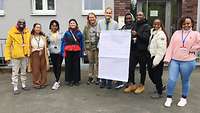 Menschen stehen in einer Gruppe zusammen und halten ein beschriebenes Blatt Papier in die Kamera.