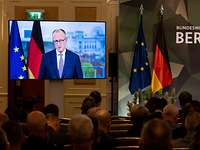 Ein Monitor, auf dem Friedrich Merz zu sehen ist, steht vor sitzendem Publikum.