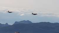 Ein A400M und eine C130 fliegen im Tiefflug in Formation über ein Schiff in einem Fjord hinweg.