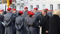 Mehrere Soldatinnen und Soldaten stehen auf einem Platz und ein Mann im schwarzen Mantel gratuliert.