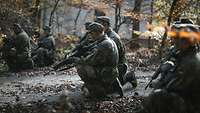 Mehrere Soldaten knien auf dem Waldboden mit Gewehr in der Hand.