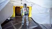 Eine Person im Schutzanzug mit der Aufschrift „EAL DEKON“ steht in einem Zelt.