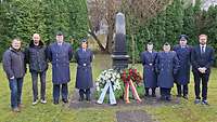 Drei Zivilisten und fünf Soldaten stehen an einer Stele mit zwei Kränzen und blicken in die Kamera.