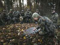 Im Wald knien mehrere Soldaten um eine Ausbilderin und erklärt etwas. 