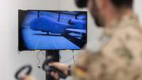 Ein Soldat mit VR-Brille und Controllern steht vor einem Monitor, auf dem eine Drohne zu sehen ist.