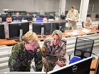 Several soldiers from different nations are sitting and standing in front of monitors in a room.