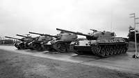 Schwarz-Weiß-Aufnahme: Vier Panzer stehen nebeneinander auf einer asphaltierten Fläche.