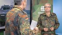 Zwei Soldaten stehen sich gegenüber und sprechen miteinander. Neben ihnen steht ein großer Monitor.