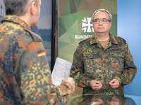 Zwei Soldaten stehen sich gegenüber und sprechen miteinander. Neben ihnen steht ein großer Monitor.