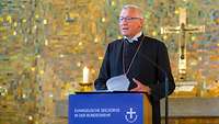 Ein Bischof steht an einem Rednerpult. Hinter ihm hängt ein Kreuz an der Wand.