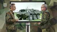 Zwei Soldaten stehen sich an einem Tisch gegenüber und sprechen. Hinter ihnen steht ein Bildschirm.