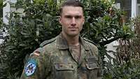 Ein Soldat steht vor einem grünen Busch und schaut in die Kamera.