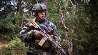 Ein Soldat steht mit Gewehr und Gefechtsanzug im Wald.