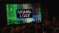 Ein großer Bildschirm mit bunten Linien zeigt den Zuschauenden die verzerrte Aufschrift Signal#en lost#en.
