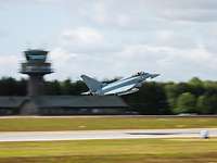 Ein Kampfjet beim Start, im Hintergrund ein Tower und Flughafeneinrichtungen