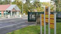 Ein grünes Schiebetor und Messingschilder, dahinter ein Wachgebäude und Schlagbäume