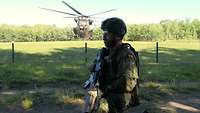 Ein bewaffneter Soldat steht neben einem Flugfeld, hinter dem Zaum landet gerade ein Hubschrauber.