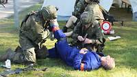 Zwei Soldaten versorgen einen verletzten Menschen in einem blauen Overall auf einer Wiese.