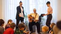 Eine Frau sitzt auf einem Stuhl und spielt Gitarre. Vor ihr sitzen Kinder auf dem Boden.