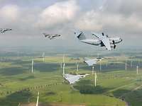 Fünf militärische Flugzeuge fliegen über einer Landschaft mit Windrädern.