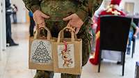 Ein Soldat hält zwei Papiertüten hinter dem Rücken. Im Hintergrund sitzt ein Nikolaus.