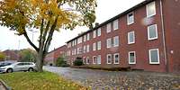 Längliches, dreistöckiges Backsteingebäude, davor Rasen, Autos und ein Baum
