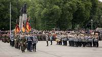 Soldaten marschieren mit Truppenfahnen über einen Platz. Eine Gruppe von Soldaten musiziert daneben.