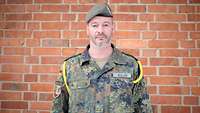 Soldat mit grau-grünem Barett vor einer Backsteinwand schaut freundlich in die Kamera.