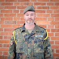 Soldat mit grau-grünem Barett vor einer Backsteinwand schaut freundlich in die Kamera.