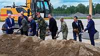 Männer in Anzug und Uniform stehen bei einem Spatenstich mit Schaufeln in der Hand vor einem Sandhaufen.