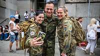 Ein Soldat und zwei Soldatinen posieren für ein Selfie. Im Hintergrund Menschen vor einem Gebäude.