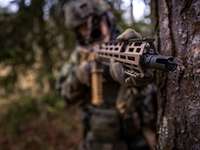 Ein Soldat steht mit seiner Waffe an einem Baum. Die Mündung der Waffe zeigt in Richtung Kamera.