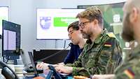 Soldaten und ein Zivilist sitzen an einem Schreibtisch vor Monitoren