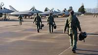 Mehrere Kampfpiloten gehen zu Kampfflugzeugen von Typ Eurofighter, die nebeneinander stehen.