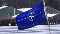 Eine NATO-Flagge steht auf einem verschneiten Platz. Im Hintergrund sind Soldaten angetreten.