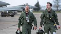 Two fighter pilots walk side by side across a runway. Fighter jets are parked in the background.