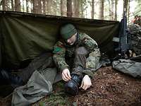 Ein Soldat sitzt hinter einer Zeltbahn im Wald und zieht sich einen Stiefel an.