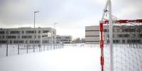 Schnee auf einem Basketball- und daneben Fußballplatz mit Toren, im Hintergrund Gebäude.