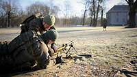 Ein Soldat liegt auf einer Matte hinter einem Maschinengewehr, einer hockt daneben und erklärt etwas
