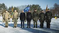 Zwei Zivilisten und fünf Soldaten stehen nebeneinander auf einem schneebedeckten Platz.