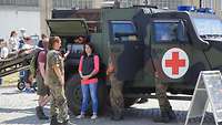 Soldaten und Zivilisten stehen an einem Militärfahrzeug mit rotem Kreuz und sprechen miteinander.
